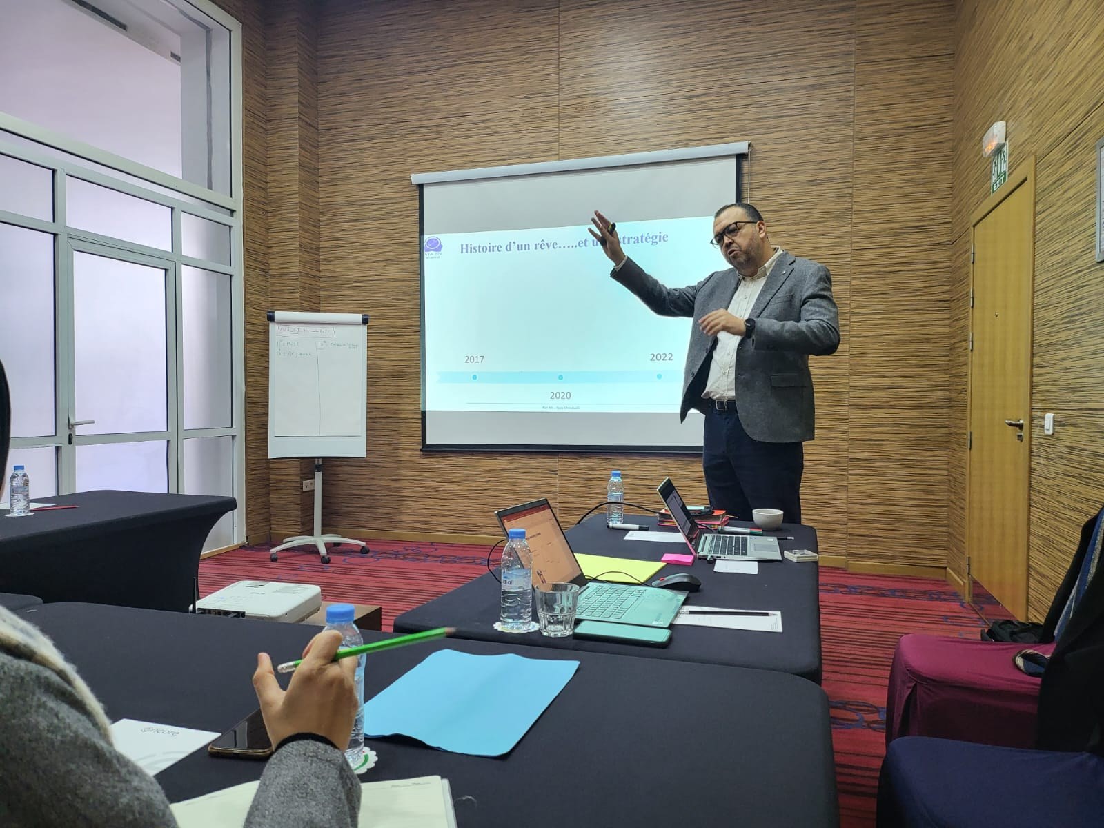 Ilyas Choubaili presenting to a seated workshop group, gesturing toward a projected slide titled “Histoire d’un rêve… et une stratégie”.